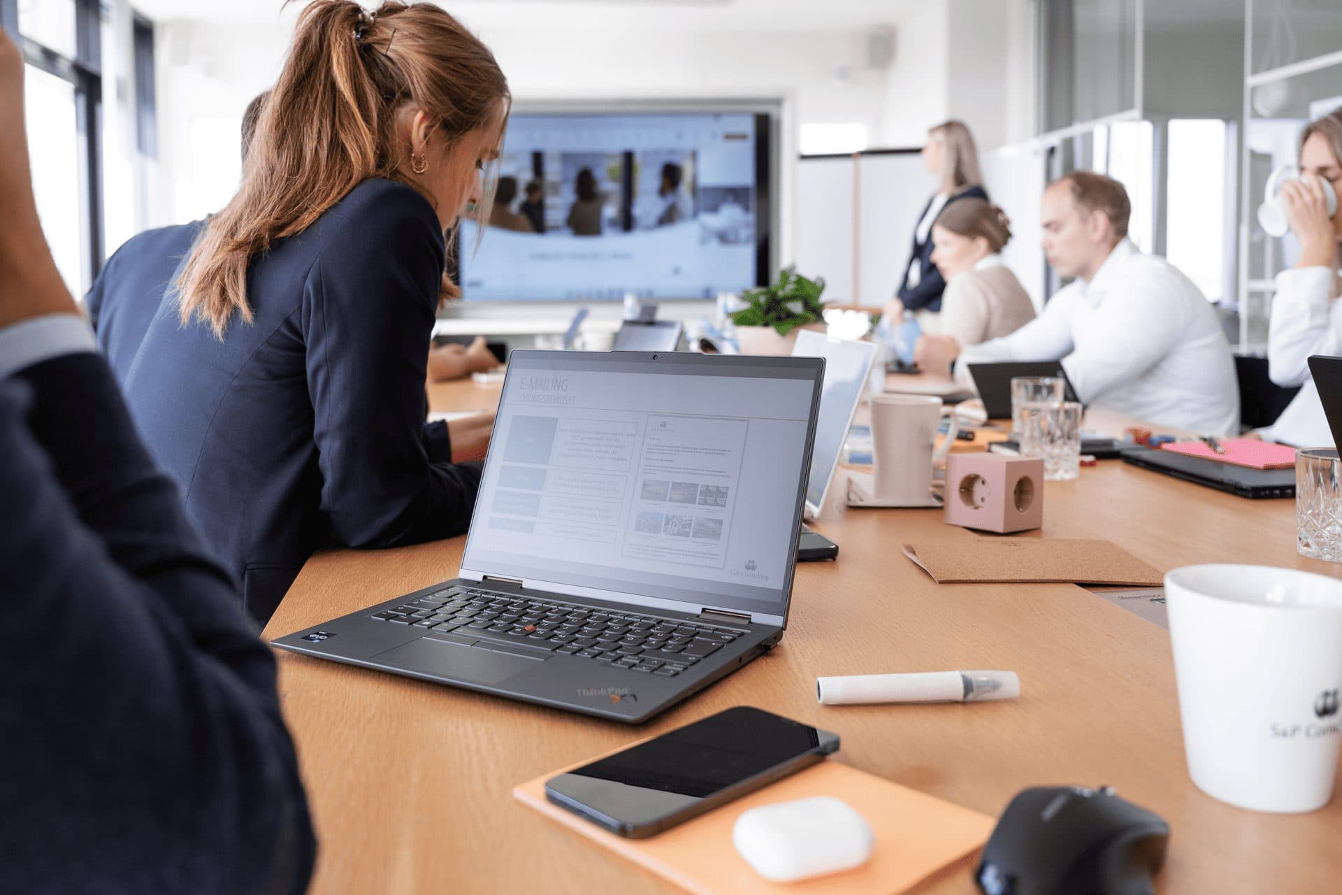 Laptop auf einem Besprechungstisch, dahinter eine Arbeitsrunde